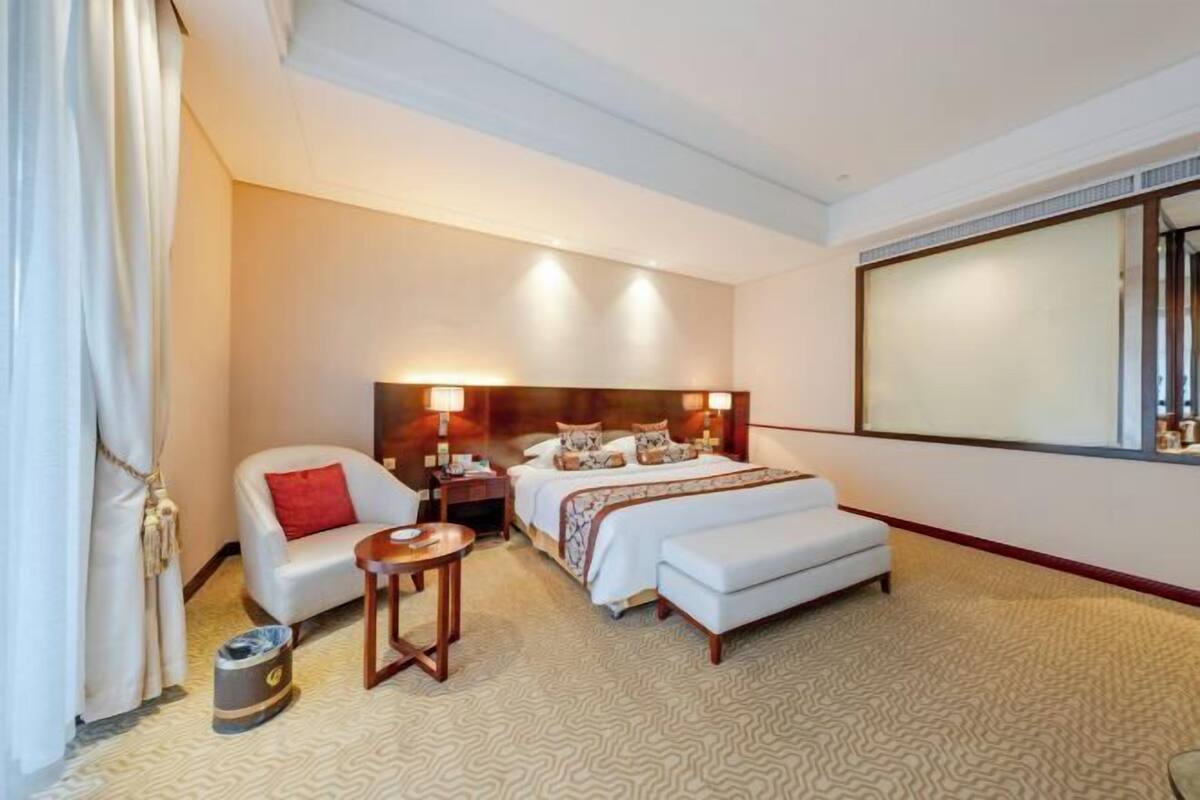 Quarto de hotel com cama, uma cadeira, mesa pequena, mesa de cabeceira e uma janela grande.