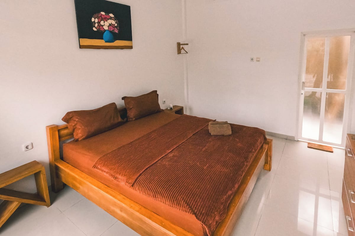 A bedroom with a wooden bed, a nightstand, a bedside table, a wall-mounted painting, and a door with a glass panel.