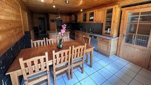 Interior em madeira com mesa e cadeiras de jantar, área de cozinha com armários e janela, e piso ladrilhado.