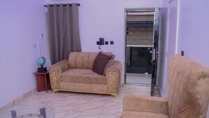 Ein Wohnzimmer mit einem beigen Sofa, einem Glastisch und einem braunen Kissen. Ein kleiner Tisch zeigt einen Globus und eine Lampe. Der Raum hat einen Fliesenboden und eine Tür, die nach draußen führt.