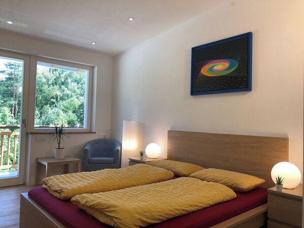 A bedroom with a double bed, a chair, a bedside table, a lamp, and a window with a view of trees.