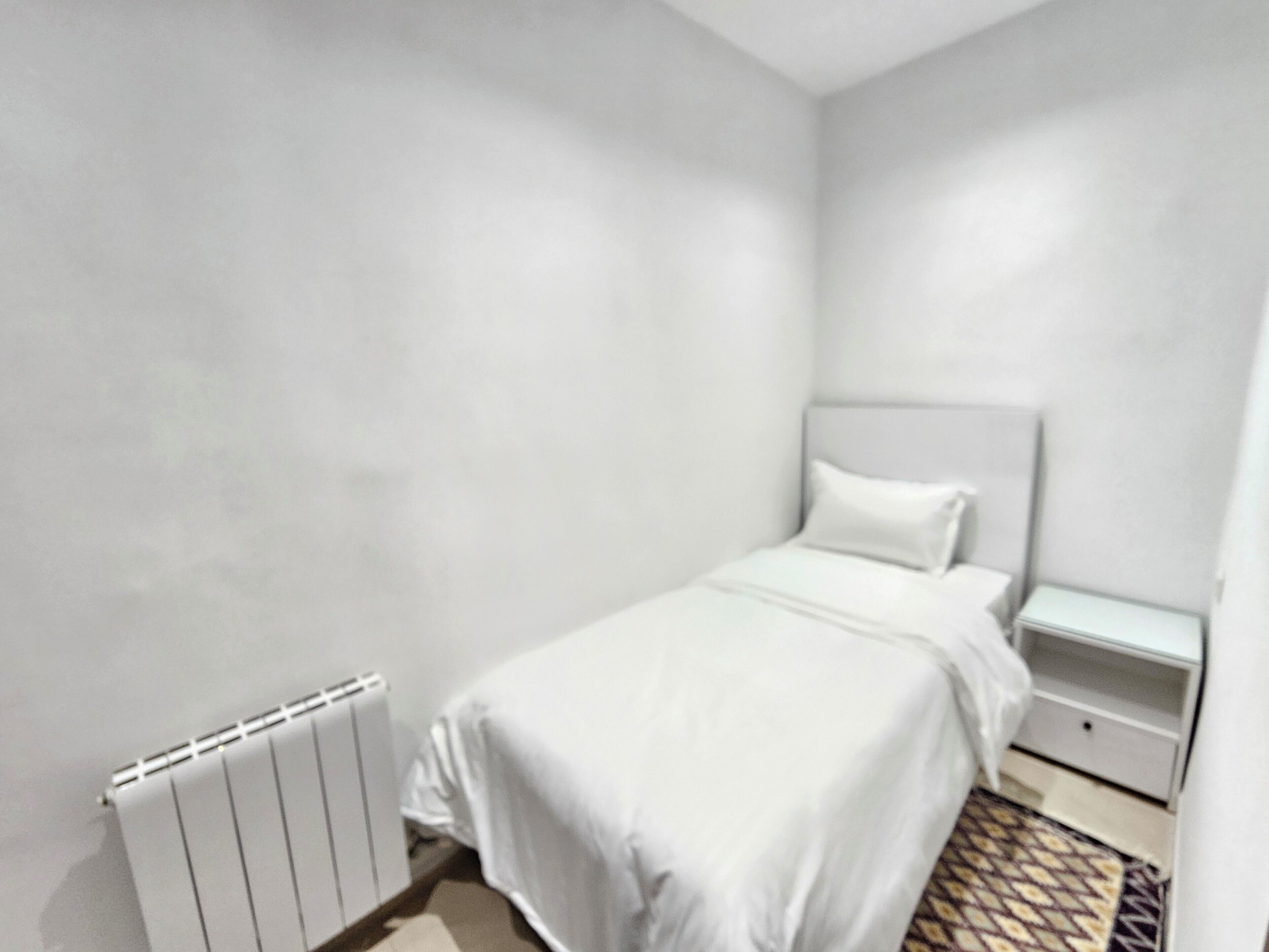 Habitación pequeña con una cama individual, radiador, mesita de noche y alfombra estampada.