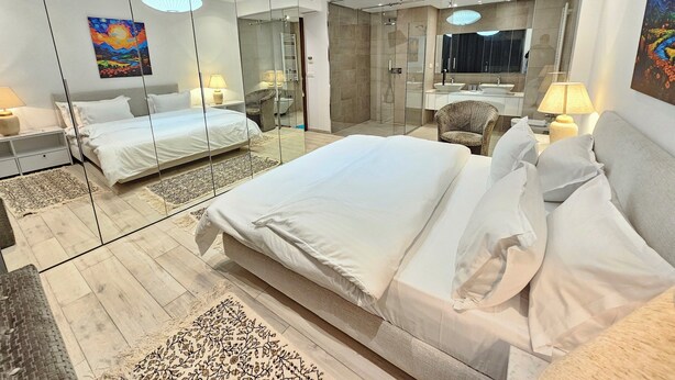 Een moderne hotelkamer met een groot bed, een zithoek met een stoel, een douchecabine van glas en een grote spiegel die de kamer weerkaatst.