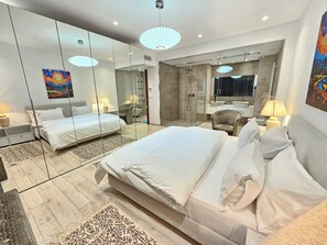 Ένα σύγχρονο δωμάτιο ξενοδοχείου με ένα μεγάλο κρεβάτι, μια μικρή γωνιά με μια πολυθρόνα, ένα ντους με γυάλινο διαχωριστικό και έναν μεγάλο καθρέφτη που αντανακλά το δωμάτιο.