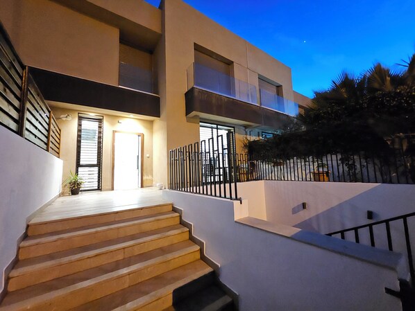 A modern building with a staircase leading to a door, surrounded by a metal fence and palm trees.