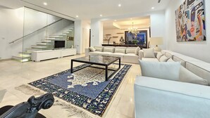 Een ruime woonkamer met een moderne bank, een salontafel en een kamerkleed. Een trap met glazen leuning loopt naar een verdieping.