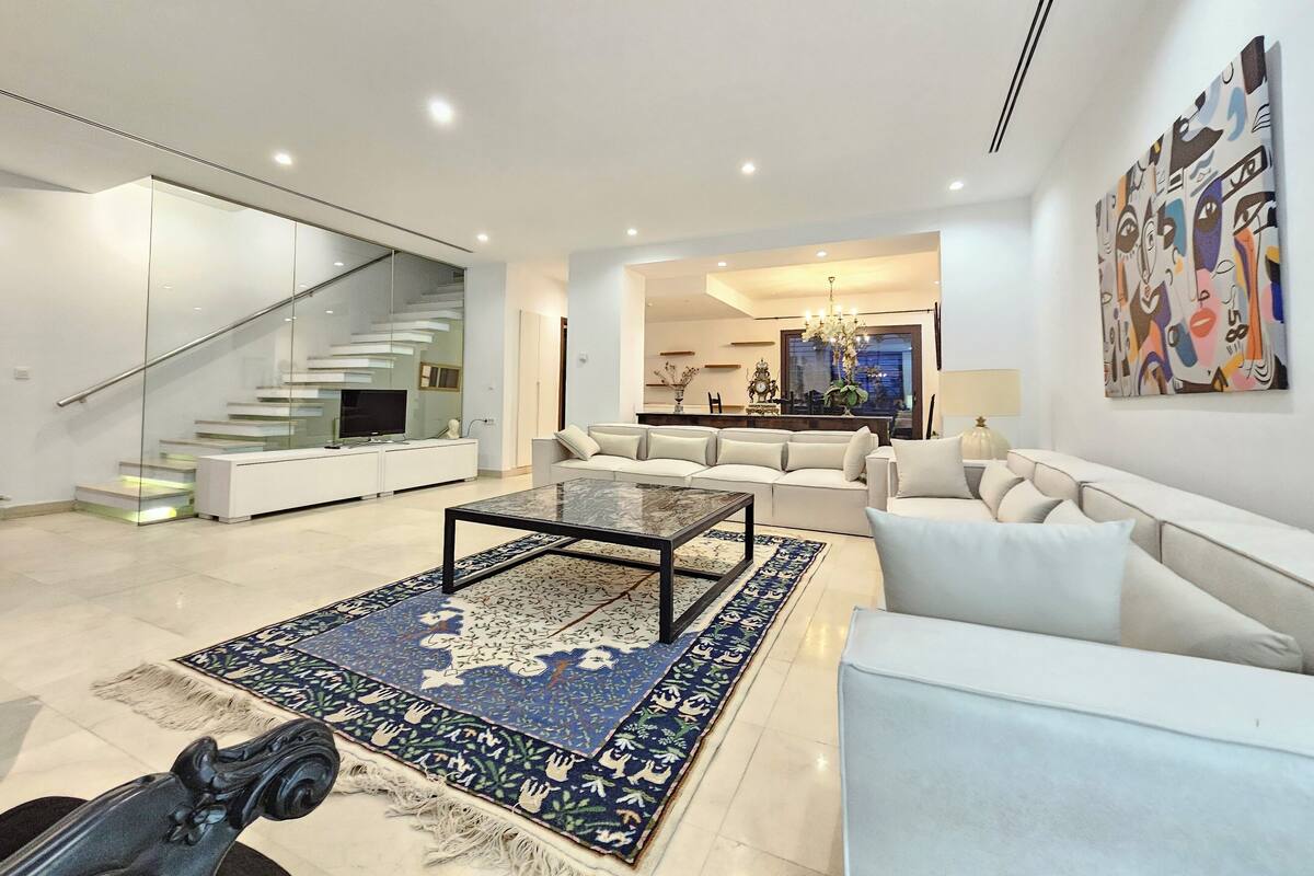 Un salón amplio con un sofá moderno, una mesa de centro y un tapete estampado. Una escalera con barandillas de vidrio conduce a un piso superior.