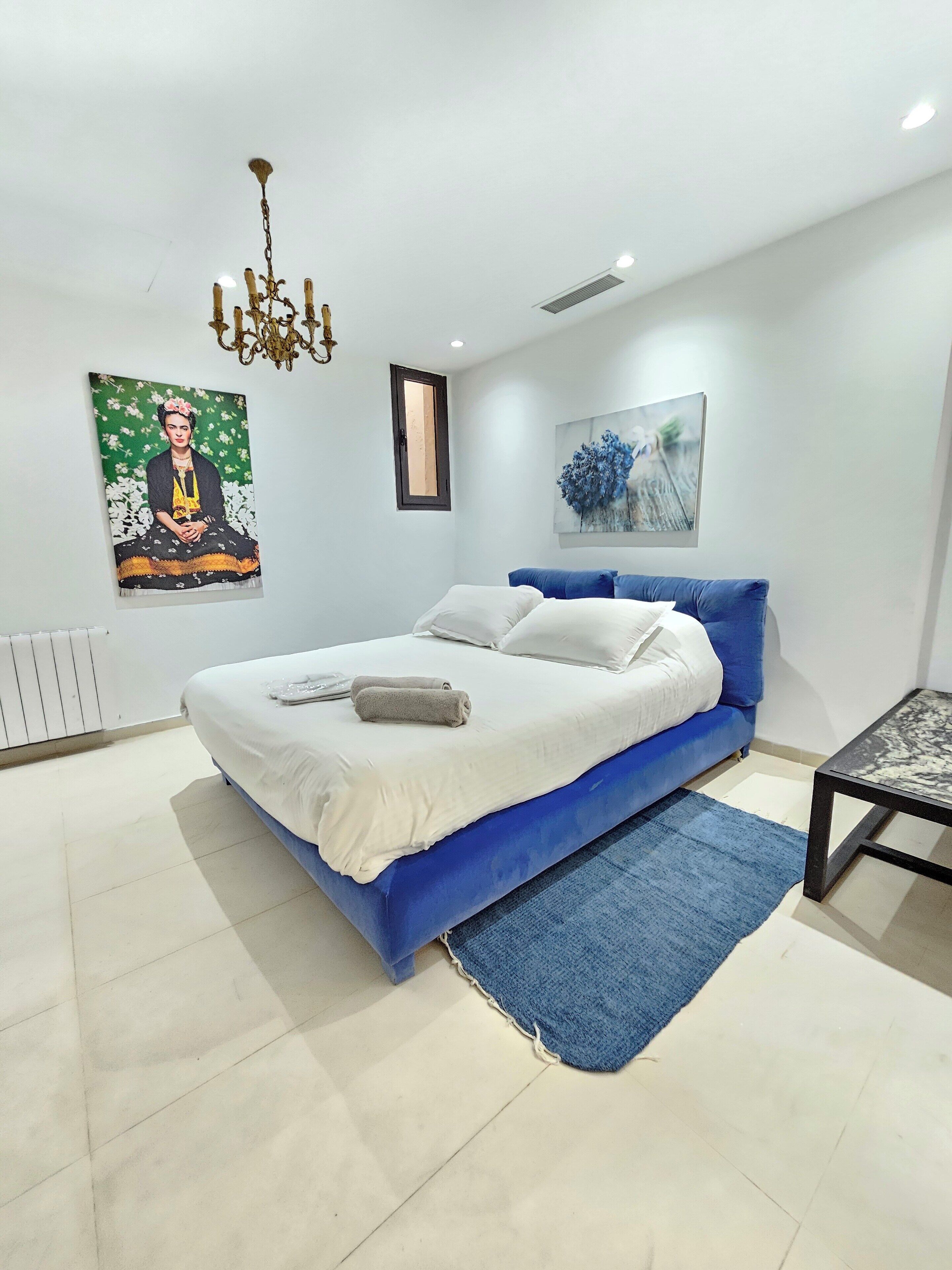 A modern bedroom with a blue bed, white bedding, a blue rug, a black table, and two wall art pieces.