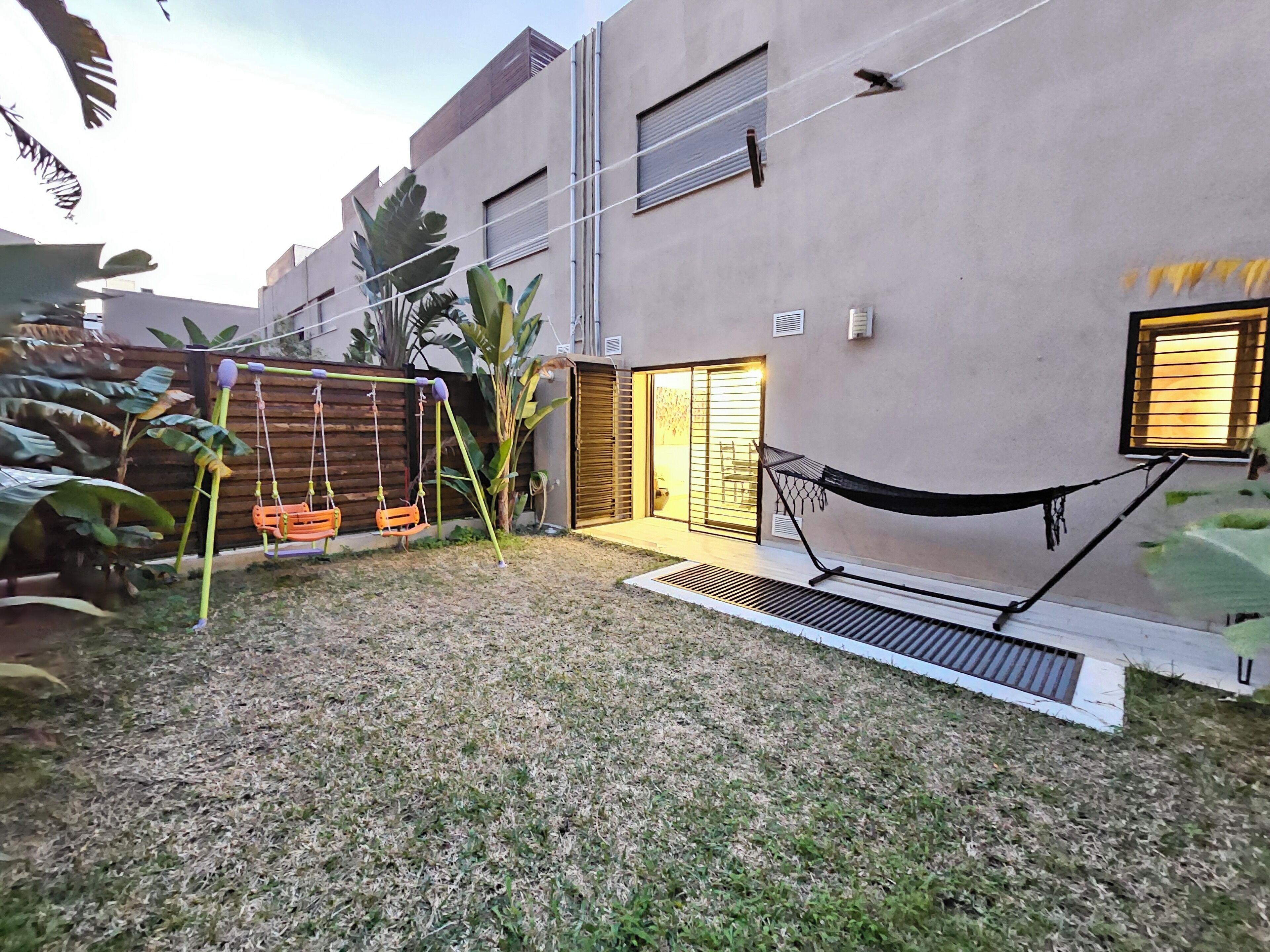 Patio trasero residencial con un columpio, una hamaca y un pequeño jardín con plantas de hojas grandes.