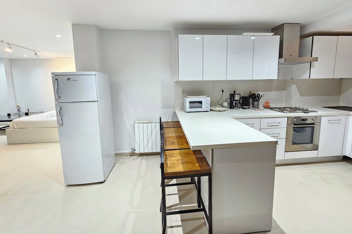 Una cocina moderna con armarios blancos, microondas integrado, cafetera y un taburete de madera.