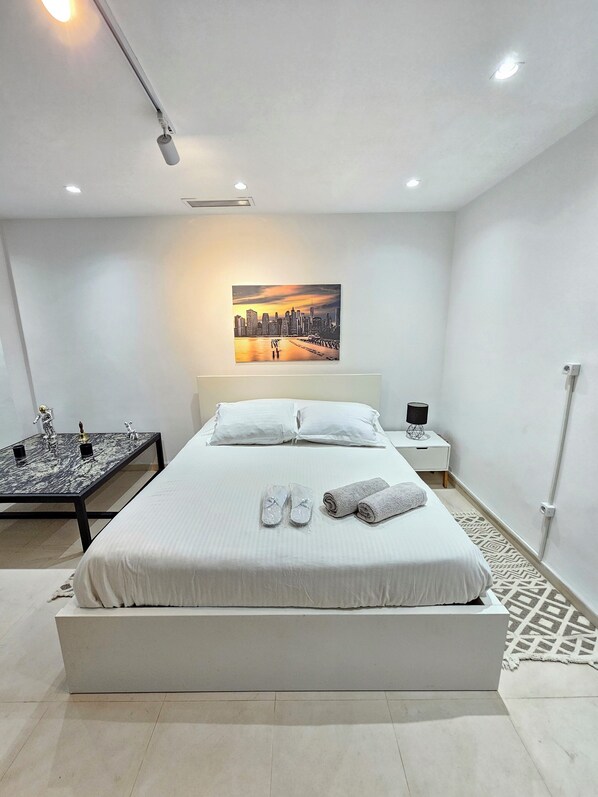 Una habitación de hotel moderna con una cama grande, un cuadro de un paisaje urbano en la pared, una mesita con figuritas y una mesita de noche con una lámpara.