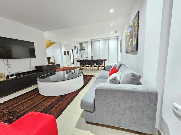A modern living room with a grey sofa, red accent chair, and a black coffee table. A flat-screen TV is mounted on the wall, and there is a dining area in the background. - Stylish Luxe House with Garden Marsa (La Marsa)