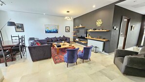 Een moderne woonkamer met een bank, salontafel en stoelen, een eethoek met tafel en stoelen, en een keuken met fornuis en planken.