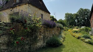 A stone house with a sloped roof, a stone wall with climbing plants, and a well-maintained garden with various shrubs and flowers.