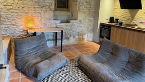 A cozy room with stone walls, a wooden table, two grey sofas, a patterned rug, and a kitchen area with a TV and coffee maker.