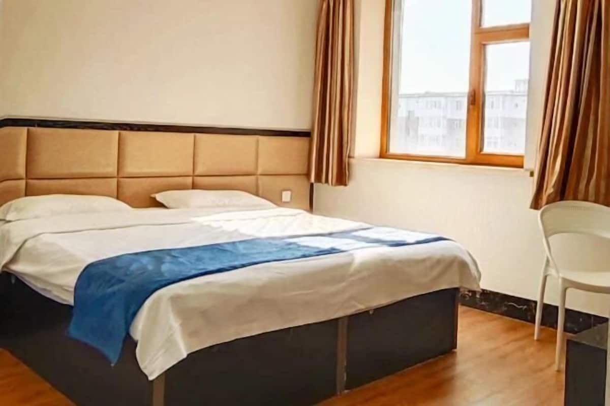 A hotel room with a bed, a chair, a window with curtains, and a view of buildings outside.