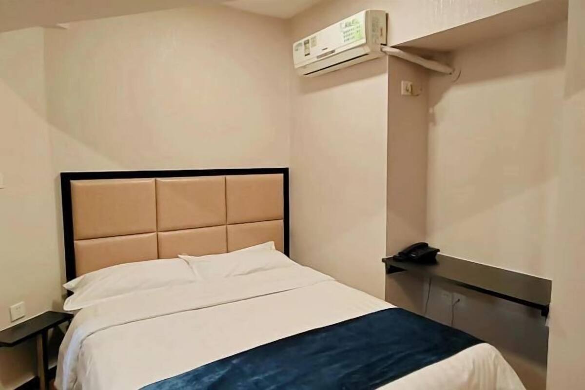 A hotel room with a bed, a bedside table, a phone, and an air conditioning unit.