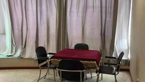 Quarto com uma mesa e quatro cadeiras, toalha de mesa vermelha e cortinas translúcidas.