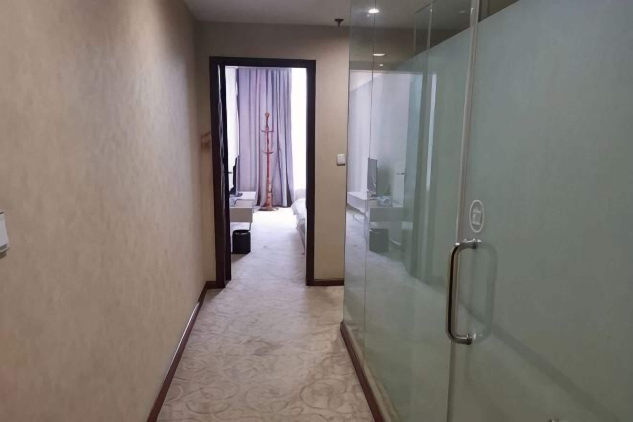 Un pasillo de hotel con piso alfombrado, una puerta de vidrio con manija, y una cama visible al fondo.