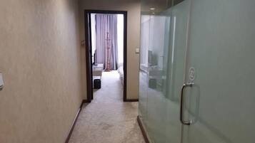Un pasillo de hotel con piso alfombrado, una puerta de vidrio con manija, y una cama visible al fondo.