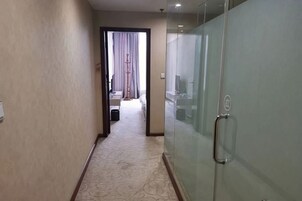 Un pasillo de hotel con piso alfombrado, una puerta de vidrio con manija, y una cama visible al fondo.