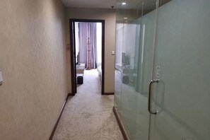 Un pasillo de hotel con piso alfombrado, una puerta de vidrio con manija, y una cama visible al fondo.