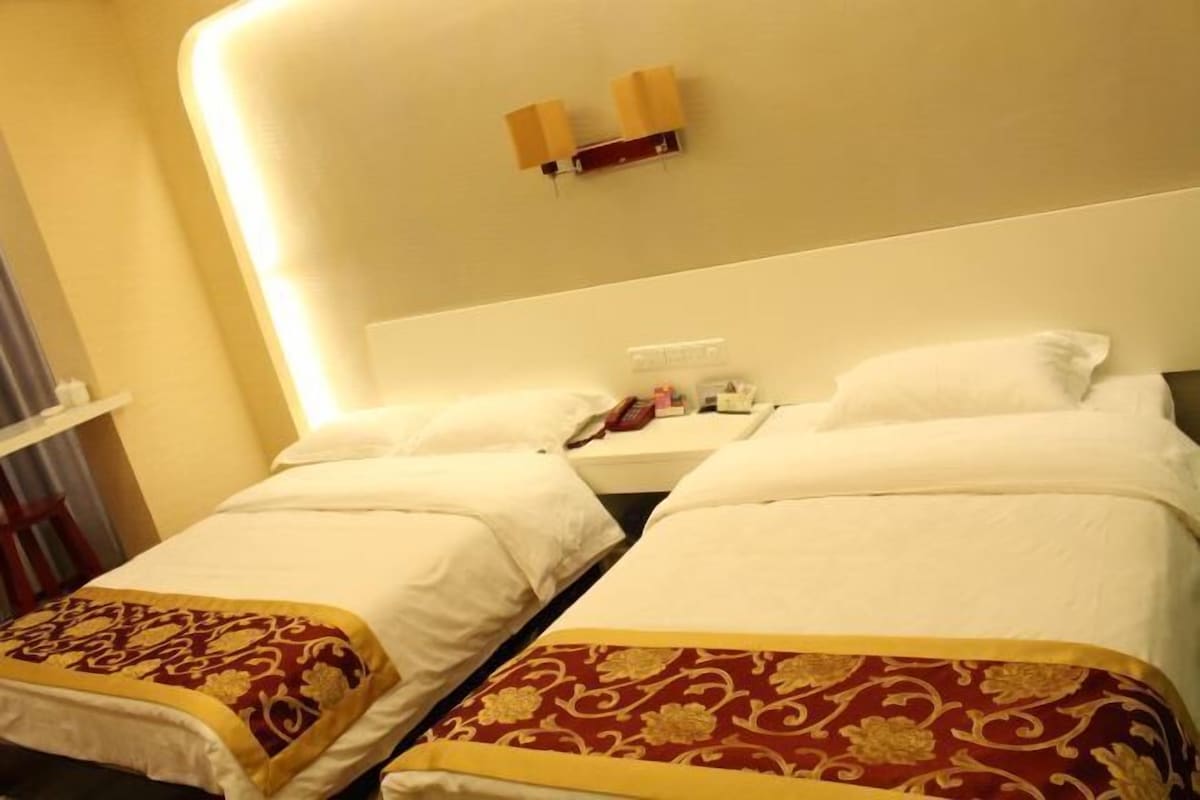 A hotel room with two beds, each with a white bedspread and a red and gold decorative pillow. There is a nightstand with a lamp and a phone.