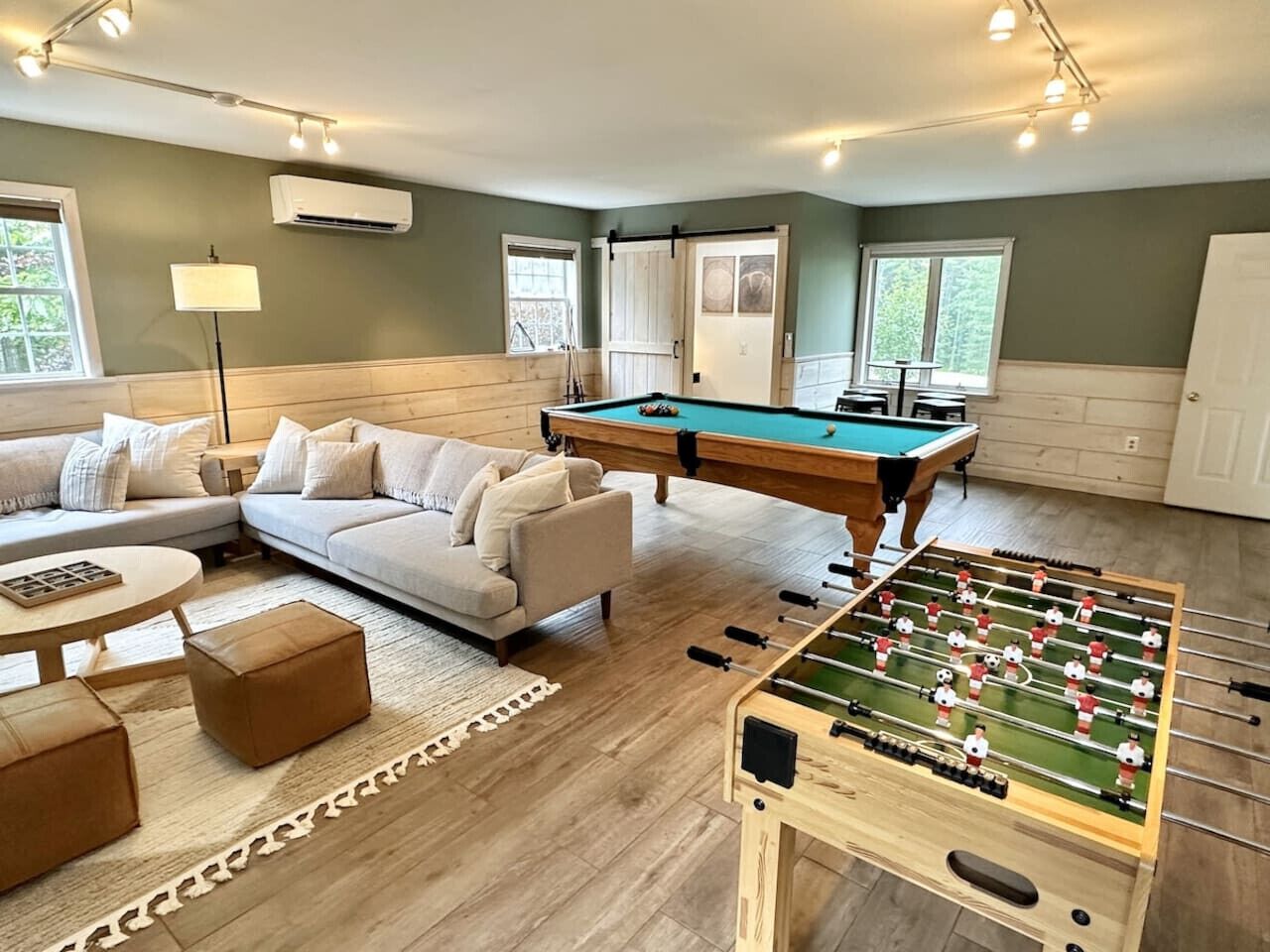 A living room with a pool table, a sofa, ottomans, and a foosball table.