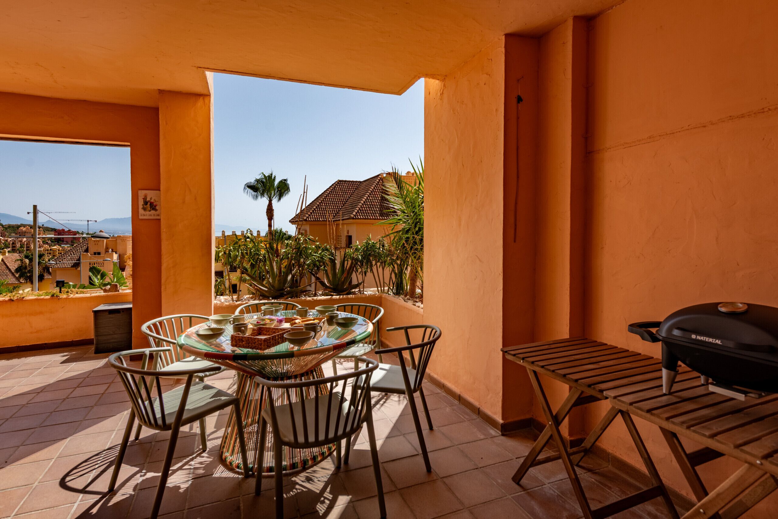 An outdoor patio with a round table and chairs, a grill, and a view of a residential area with palm trees and a house.