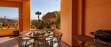 An outdoor patio with a round table and chairs, a grill, and a view of a residential area with palm trees and a house.