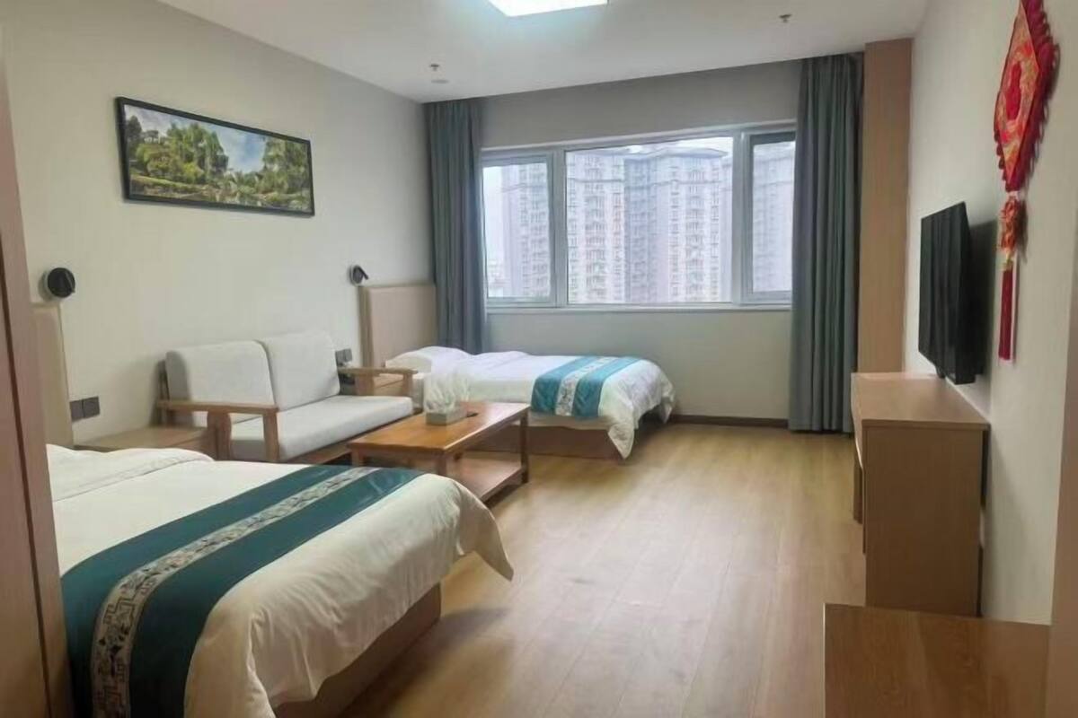 Hotellihuone, jossa on kaksi sänkyä, sohva, työpöytä, televisio ja ikkuna, josta avautuu näkymä kaupunkiin.