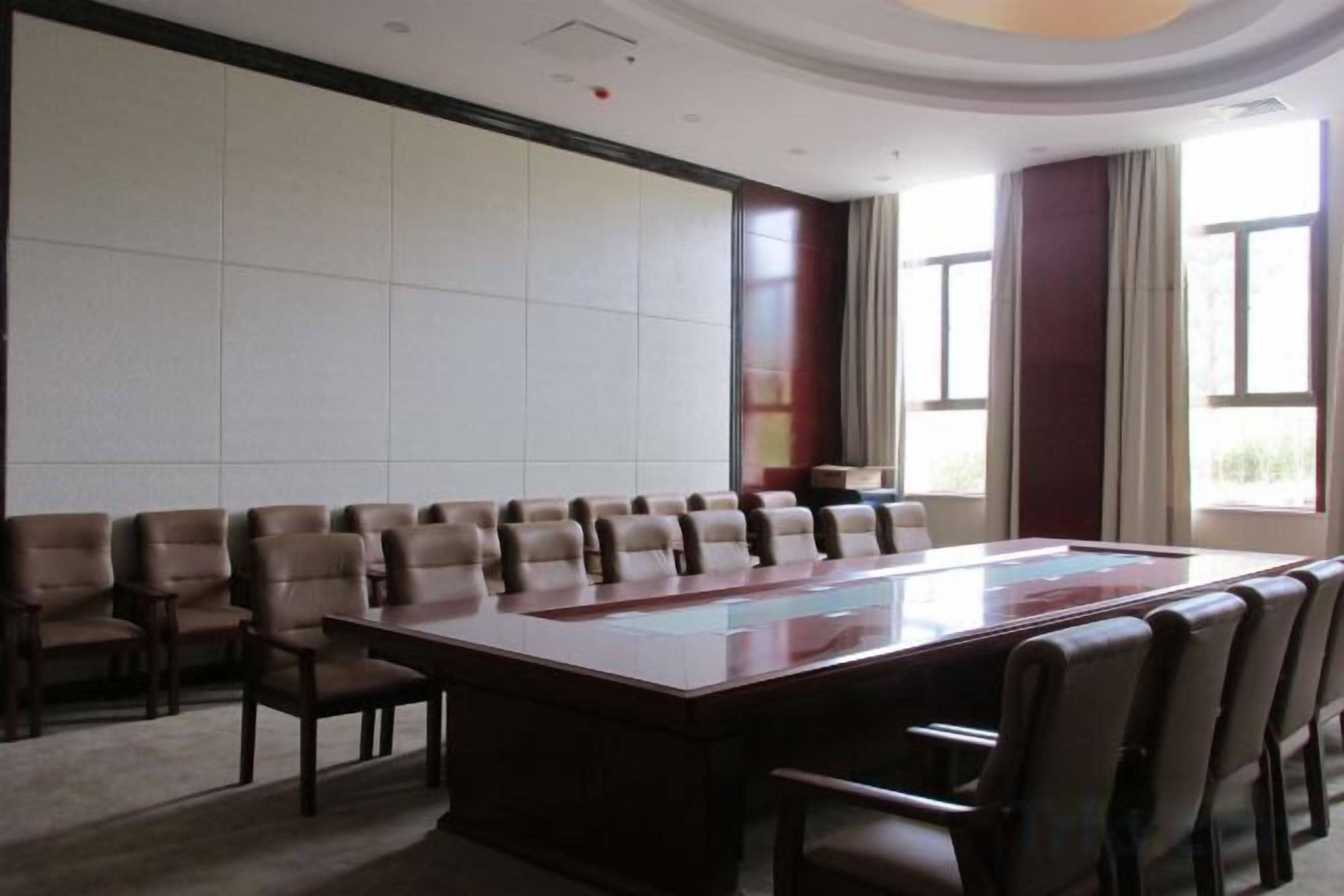 A conference room with a large rectangular table surrounded by chairs, large windows with curtains, and a carpeted floor.