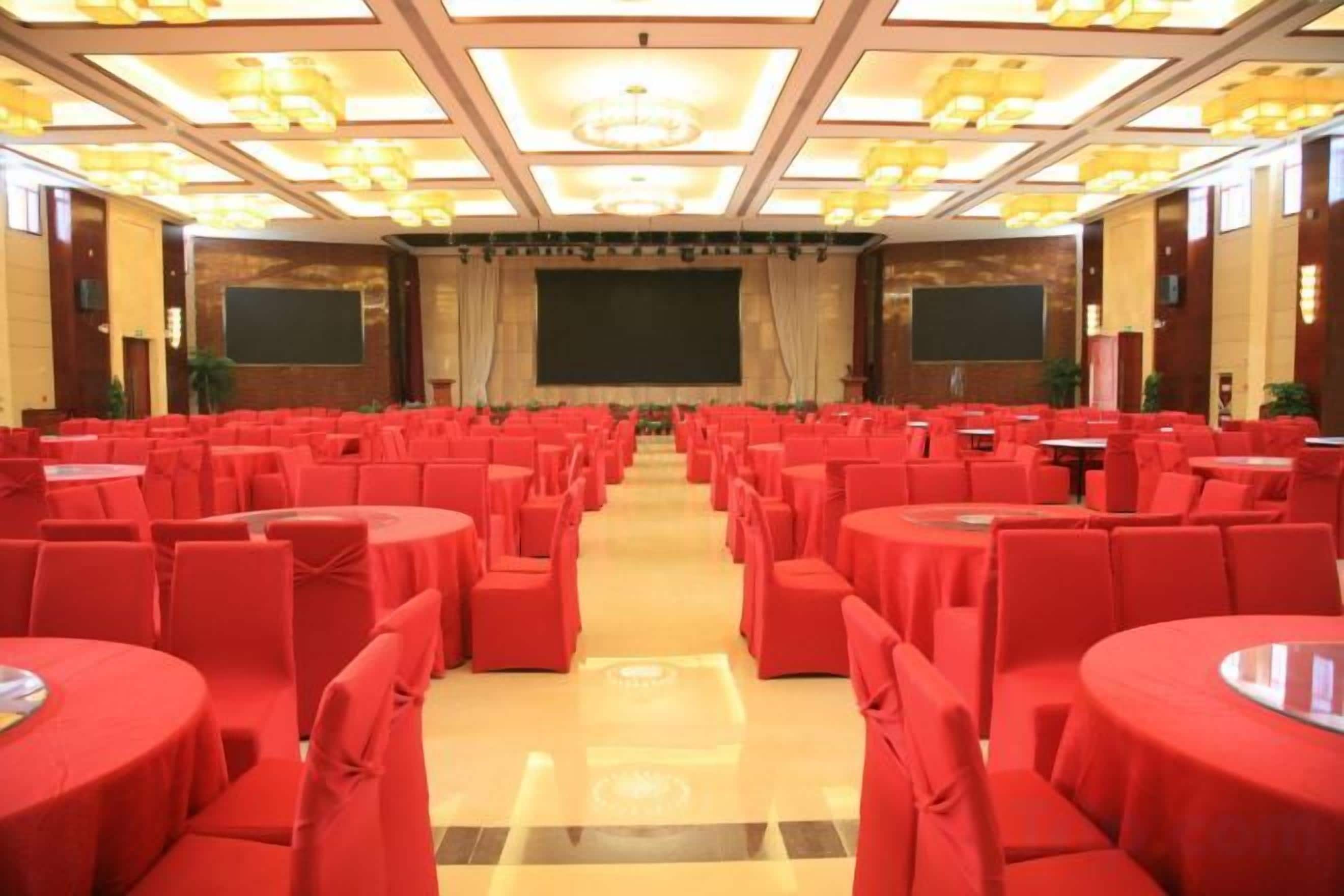A conference room with rows of tables and chairs, a stage with a screen, and a chandelier.
