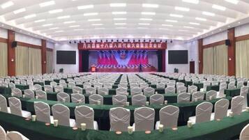 A conference room with rows of tables and chairs, a stage with a red backdrop, and a large screen.