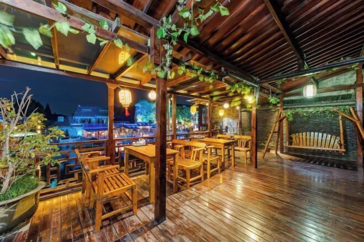 Um deck de madeira ao ar livre com mesas e cadeiras, uma área coberta com plantas penduradas e vista para edifícios tradicionais ao fundo.
