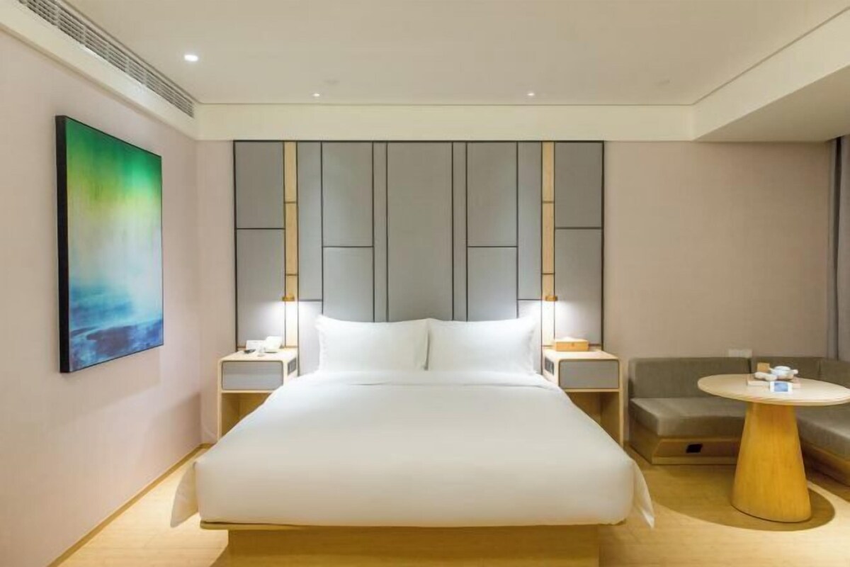 A modern hotel room with a large bed, bedside tables, a round wooden side table, and a painting on the wall.