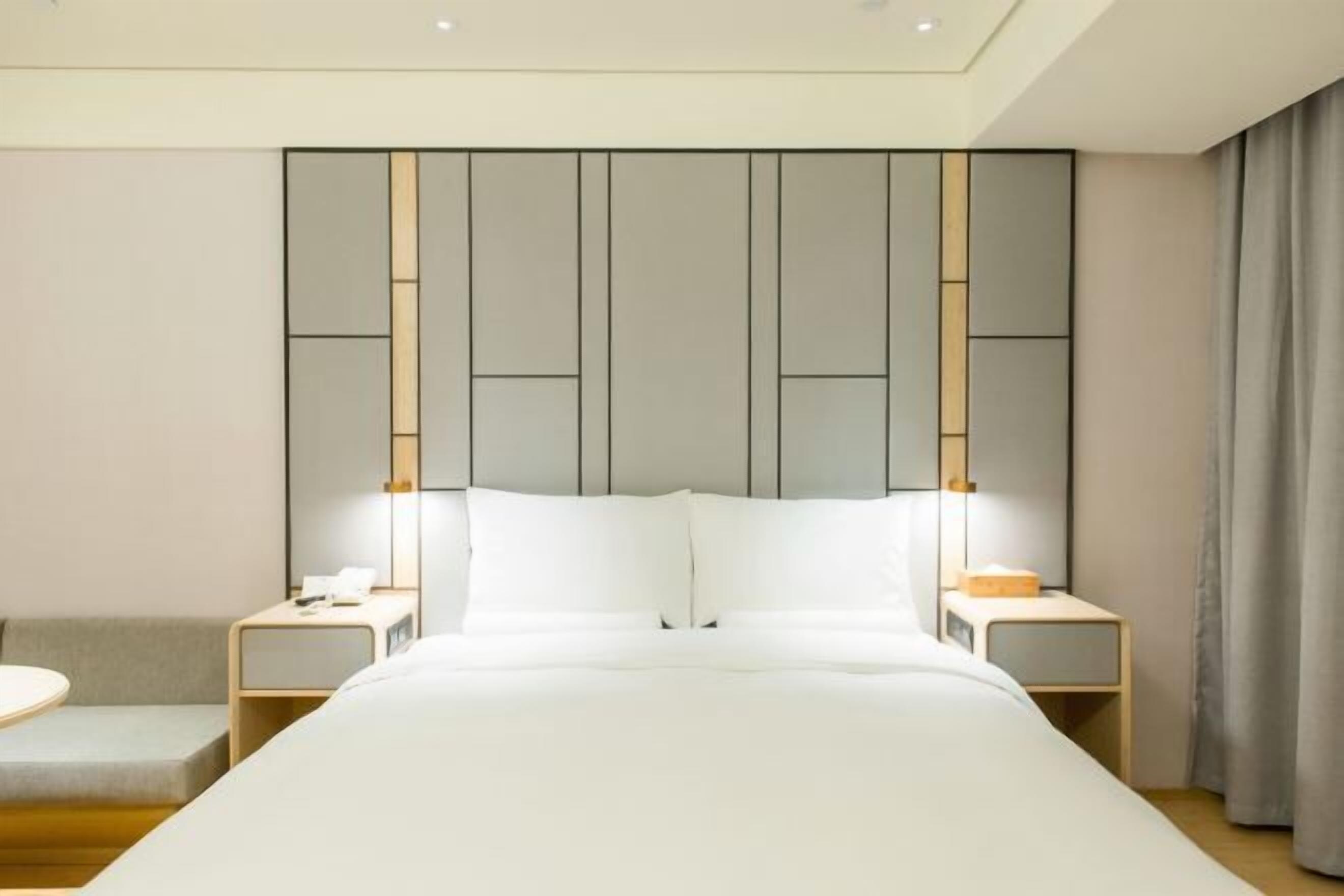 Quarto de hotel moderno com uma cama grande, duas mesinhas de cabeceira, um pequeno sofá e cabeceira com painéis verticais.