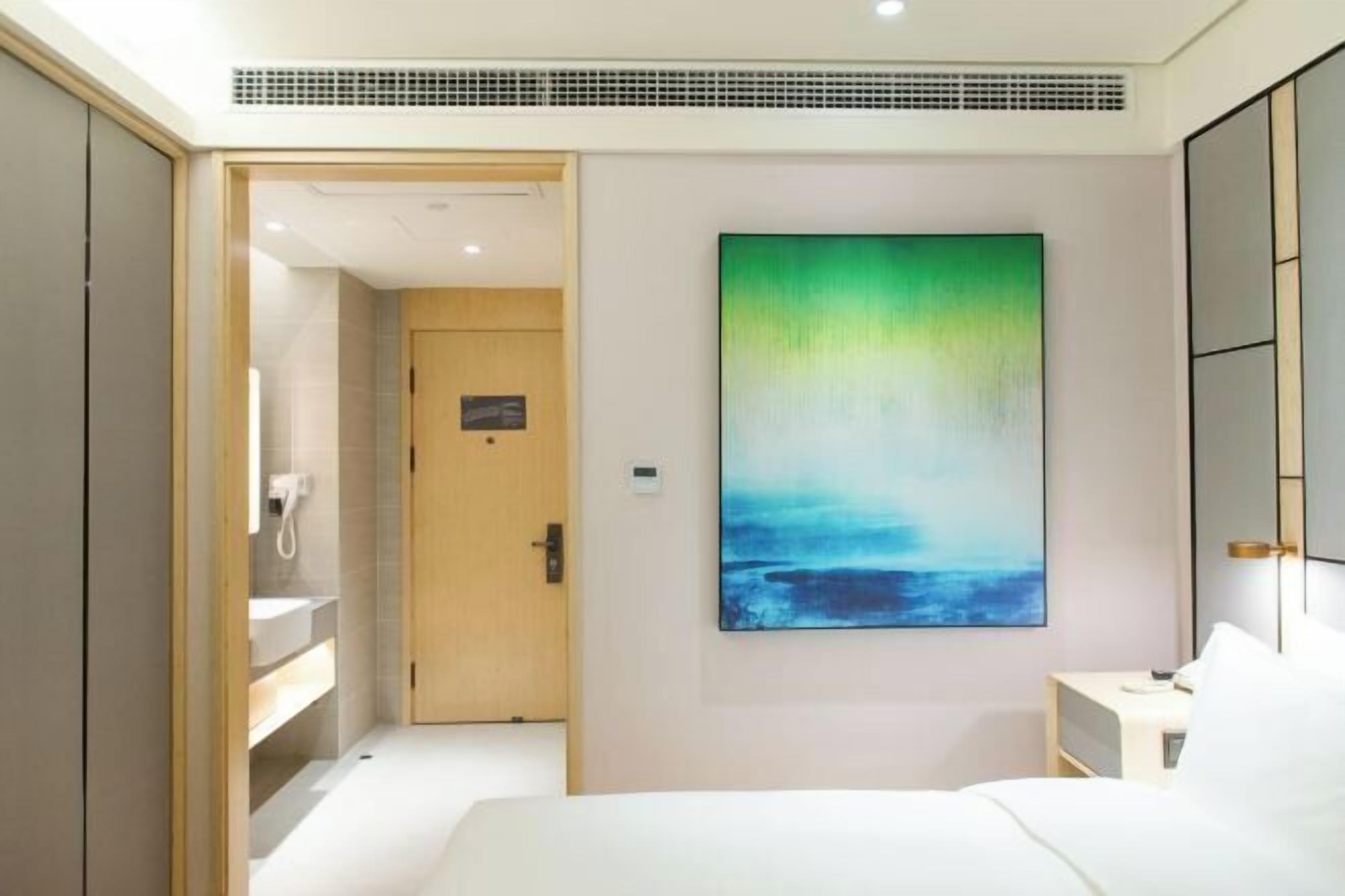A modern hotel room with a large bed, a bedside table, a wall-mounted painting, and a bathroom visible through an open door.