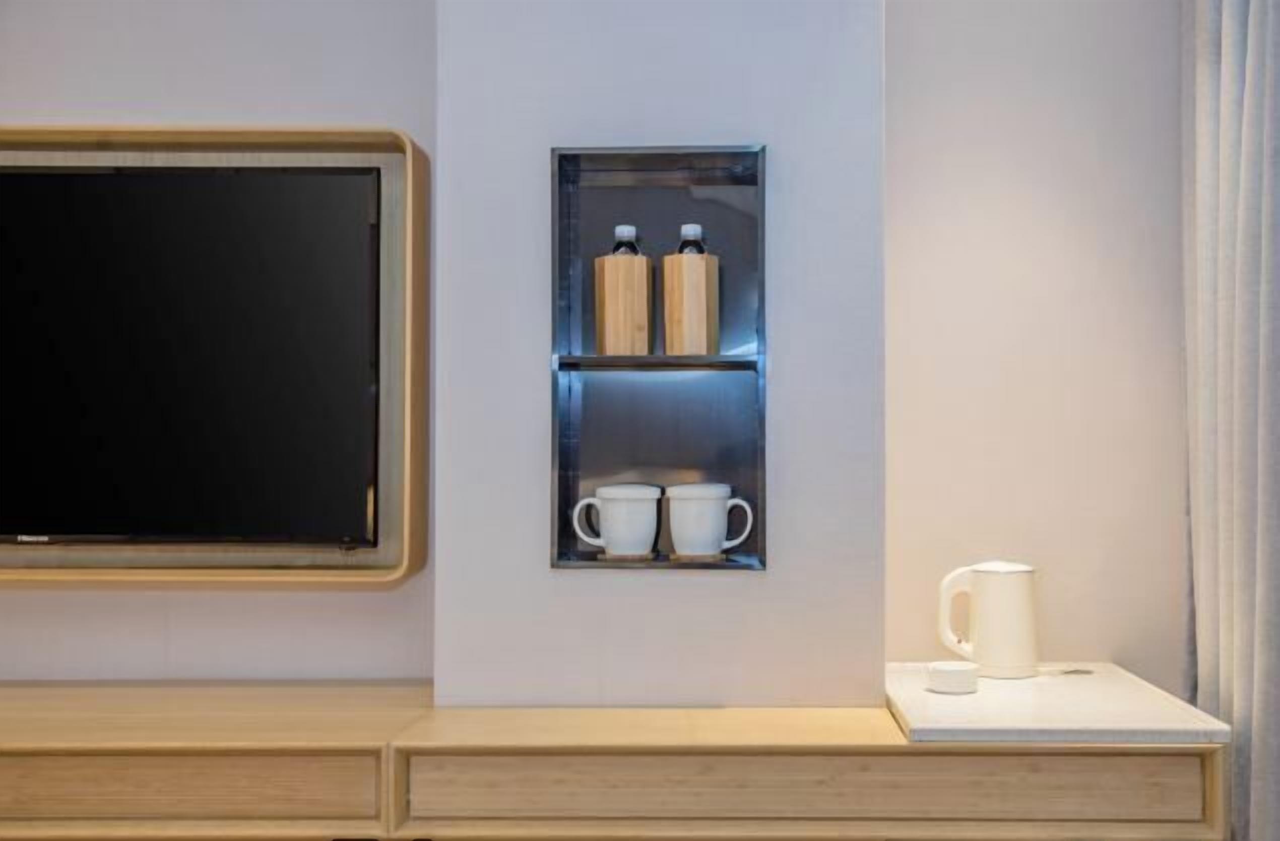 A modern hotel room with a wooden cabinet, a built-in shelf with two bottles and two cups, and a coffee maker on the counter.