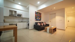 A modern kitchen with a microwave, stove, and sink. There is a black sofa with colorful pillows and a wooden coffee table. The room has white walls and a tiled floor. - suncity Apartments (Accra)