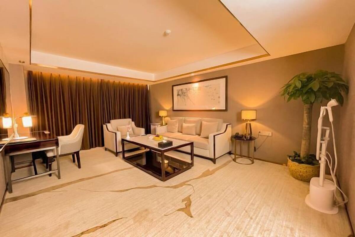 A modern hotel room with a sofa, armchairs, a coffee table, a desk, and a lamp.
