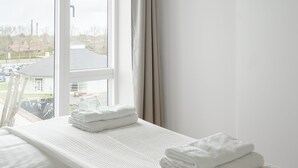 A hotel room with a large window, a bed with white linens, and a view of a parking lot and buildings outside. - Top-class 1-bedroom Apartment in Odense (Odense)