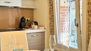 Una cocina con cafetera, hervidor y un plato de comida, un comedor con una silla y copas de vino, y una pared de ladrillo con una puerta.