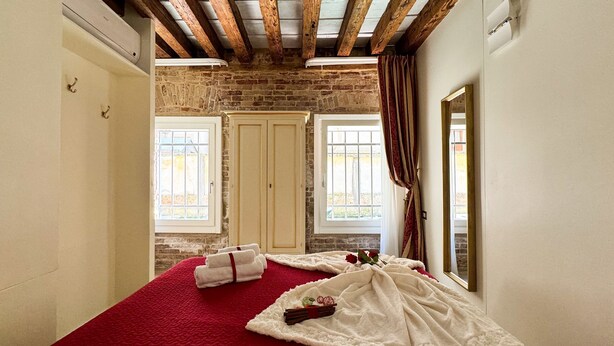 Una habitación con alfombra roja, dos ventanas con cortinas, una pared de ladrillo y techo de madera.