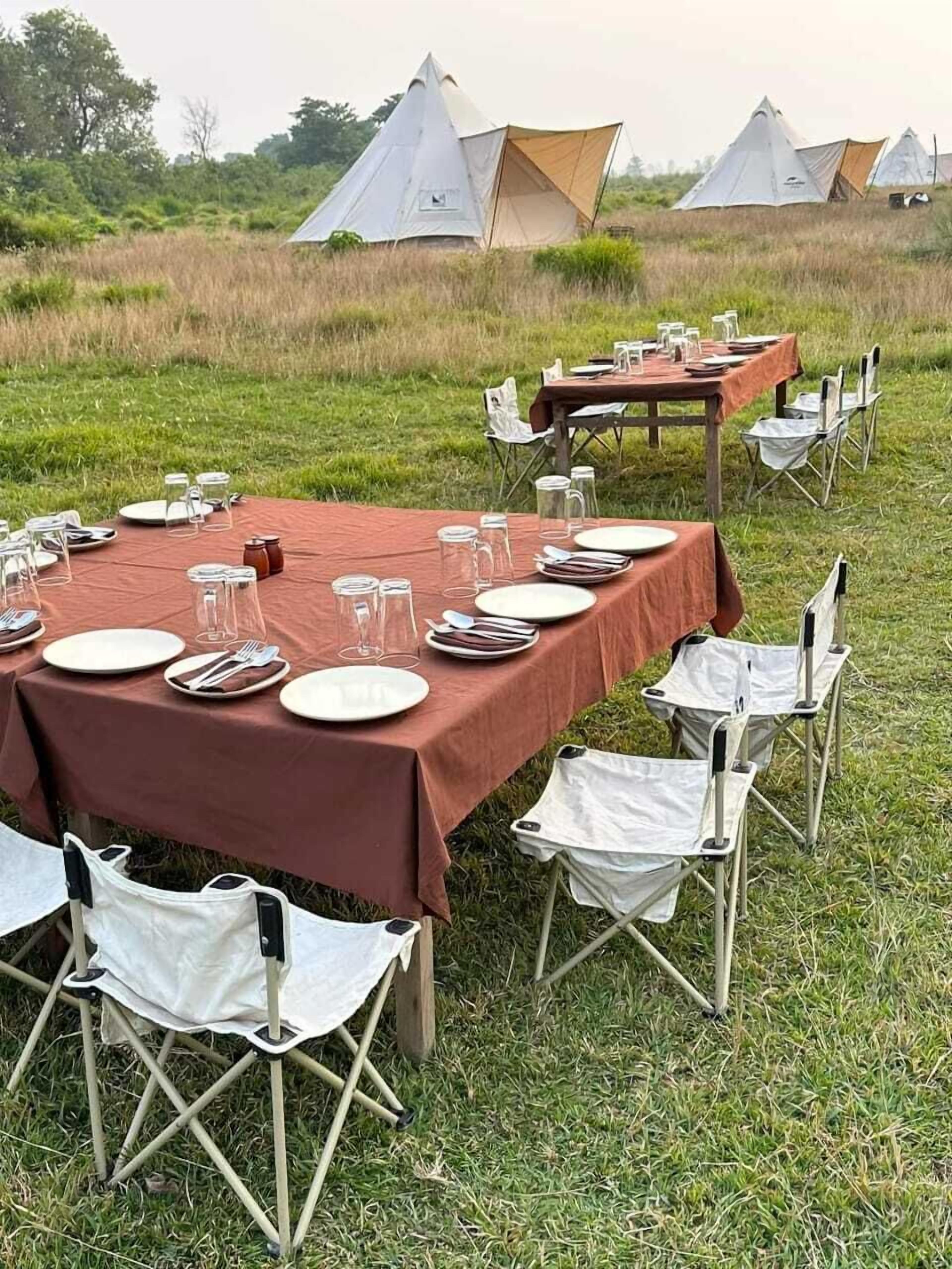 An outdoor dining setup with tables set for meals, surrounded by tents in a grassy field.