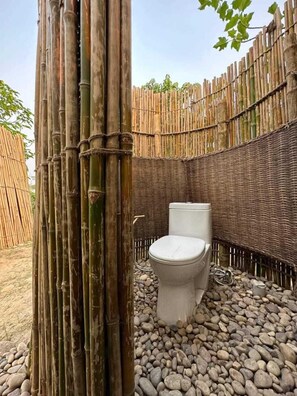 Un inodoro en un baño al aire libre con paredes de bambú y suelo de piedra.