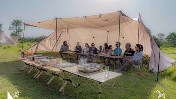 A group of people seated around a table with camping gear in a tented area.