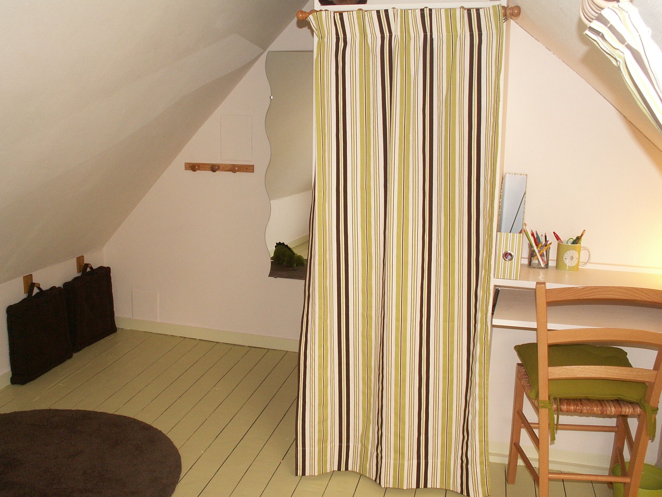A room with a sloped ceiling, a striped curtain, a wooden chair, and a desk with a mug and pens.