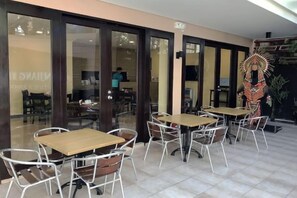 Een terras met tafels en stoelen, een decoratief wandstuk, en uitzicht op het interieur door glazen deuren.