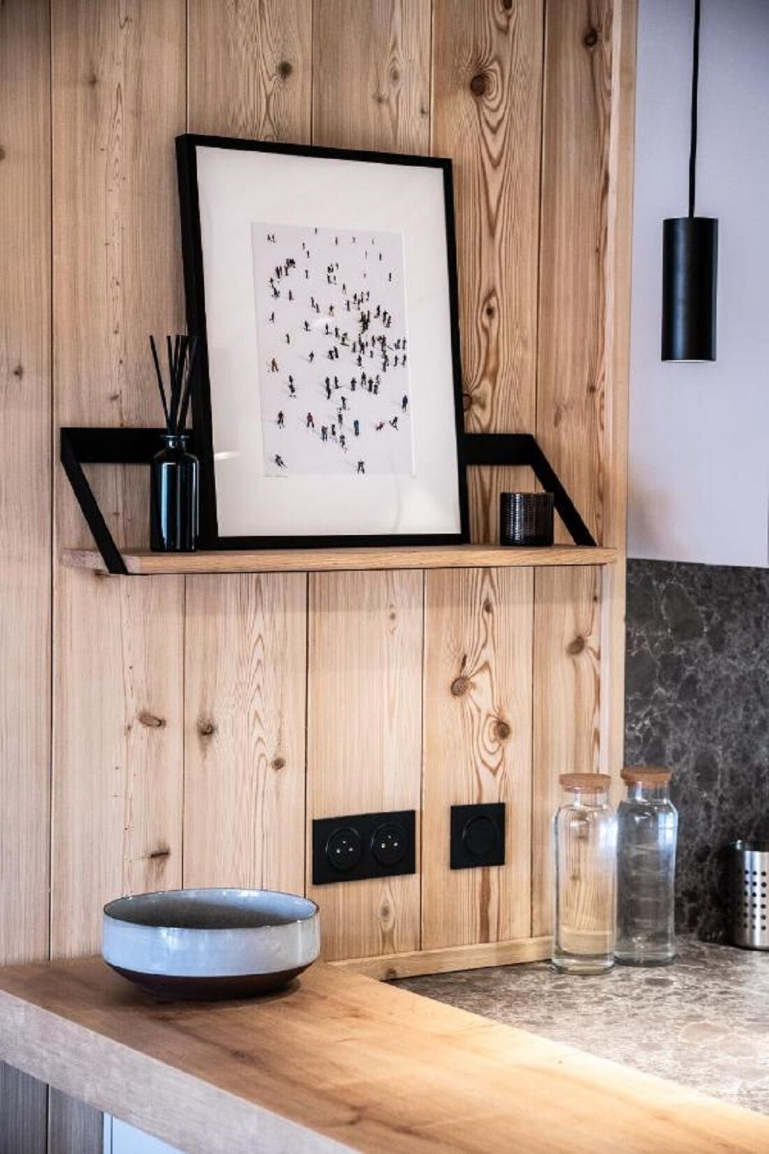 A kitchen with a wooden wall, a framed picture, a black shelf, a bowl, and some jars.
